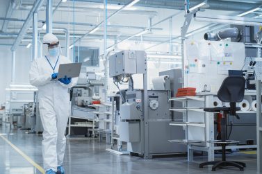 Heavy Industry Manufacturing Factory: Scientist in Sterile Coverall Walking with Laptop Computer, Examining Industrial CNC Machine Settings and Configuring Production Functionality.